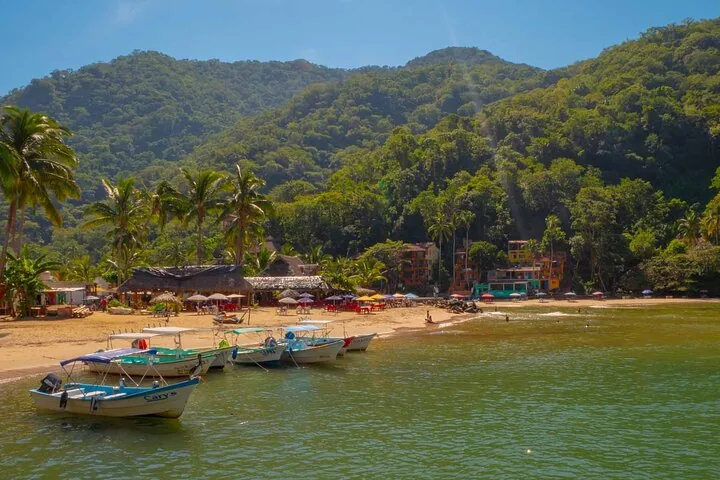 Boats moored at a sandy beach with thatched roofs and lush green hills